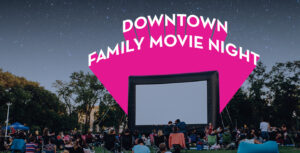 Downtown family movie night, people sitting on the grass in front of a large outdoor movie screen.
