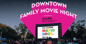 Downtown family movie night, people sitting on the grass in front of a large outdoor movie screen promoting the Super Mario Brothers movie.